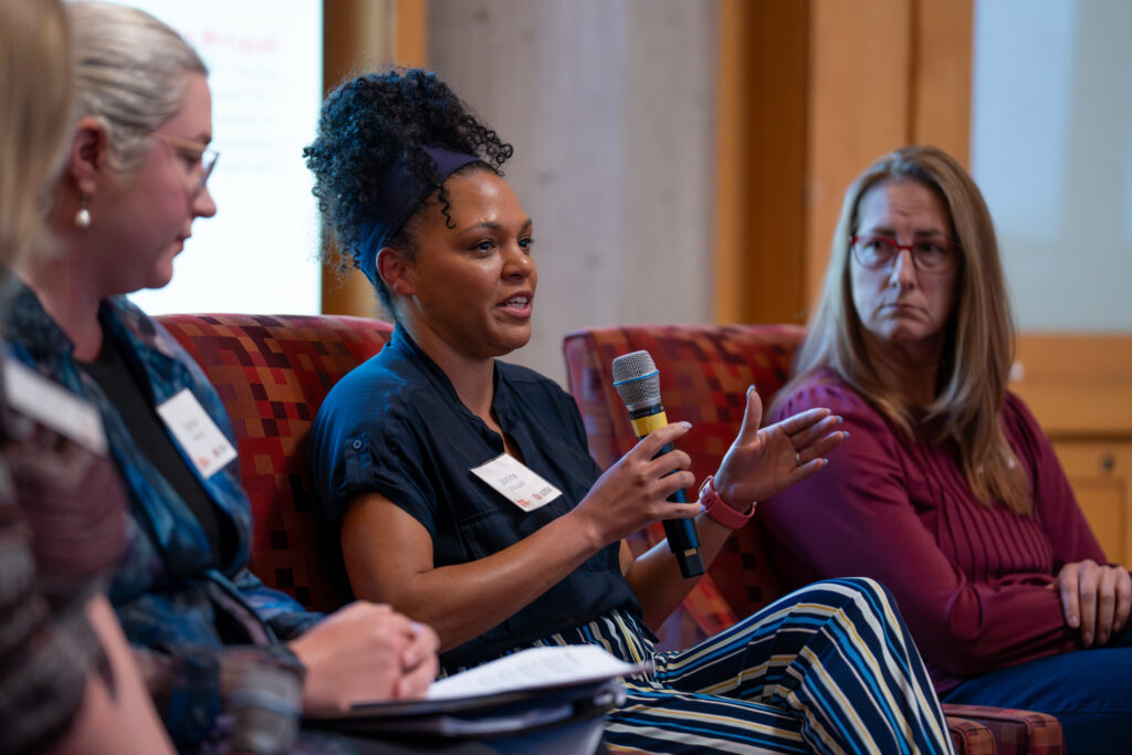 Jasmine Motupalli, founder of The Data Love Co., speaks into a microphone during the "Women Engineering AI Solutions That Matter" panel at the 2026 Bridges to Tomorrow event, seated alongside fellow panelists with their bios displayed on screen behind them.