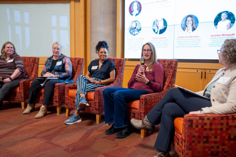 Panel of five women seated in red patterned chairs during a discussion, with a large screen displaying bios in the background.